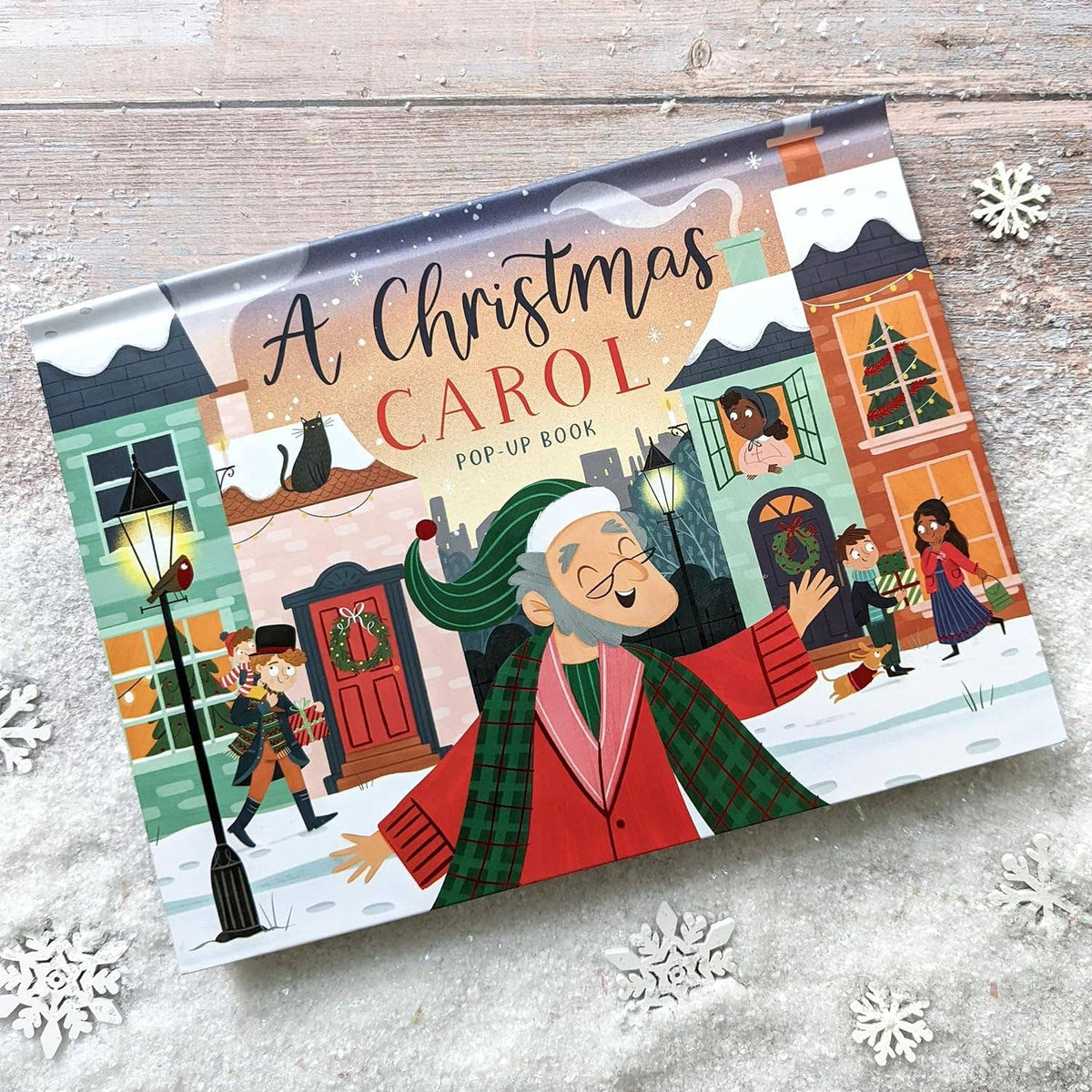 A Christmas Carol children's pop-up book showing its illustrated cover on a rustic wooden surface with decorative snowflakes