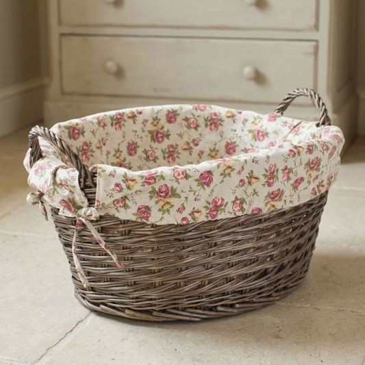 Antique Wash Willow Laundry Basket with Garden Rose Lining showing its pink floral fabric, oval wicker shape and woven handles in a country cottage bedroom.