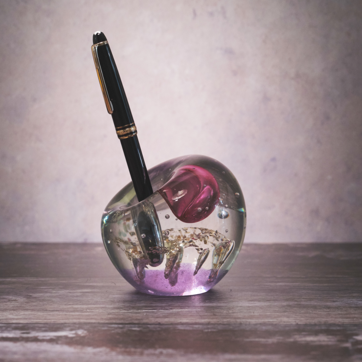 Pen Holder Glass Paperweight holding a black pen on a wooden writing desk with pink and gold-toned glass detailing