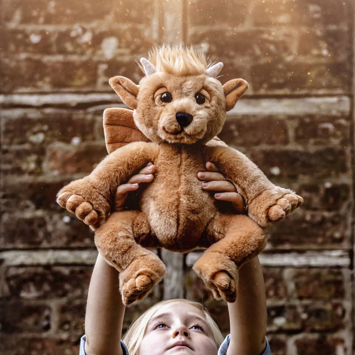 Charlie Bears Bartizan golden-brown plush gargoyle being held up by a child against a rustic brick wall