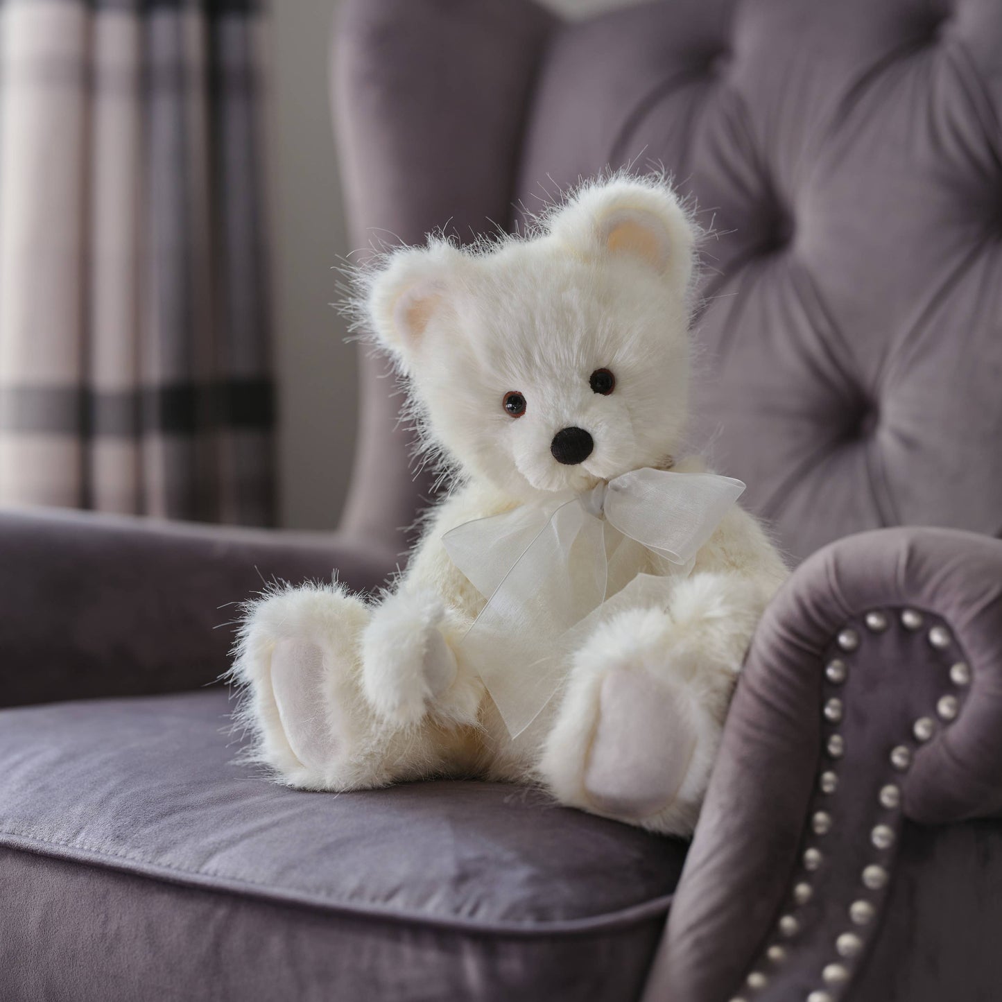 Charlie Bears Little Bean white plush teddy bear with organza bow displayed on an upholstered chair