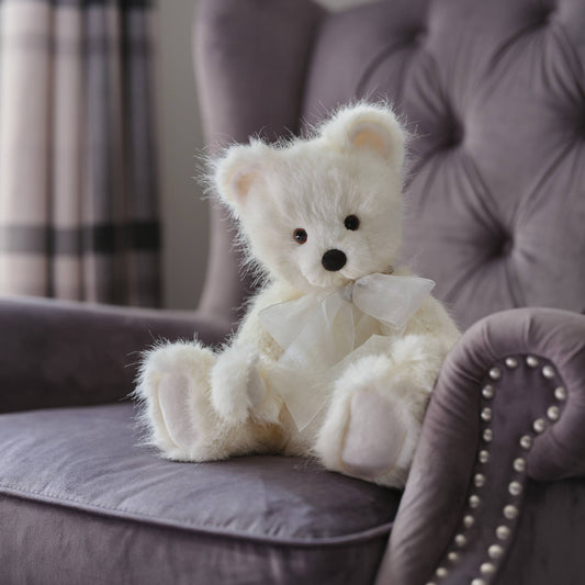 Charlie Bears Little Bean white plush teddy bear with organza bow displayed on an upholstered chair