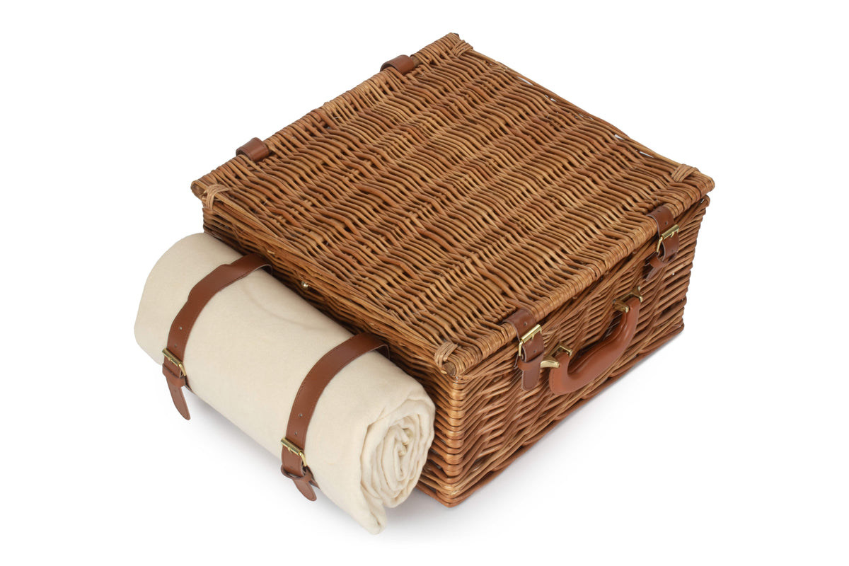Traditional rectangular willow picnic hamper closed with tan straps and attached cream picnic rug