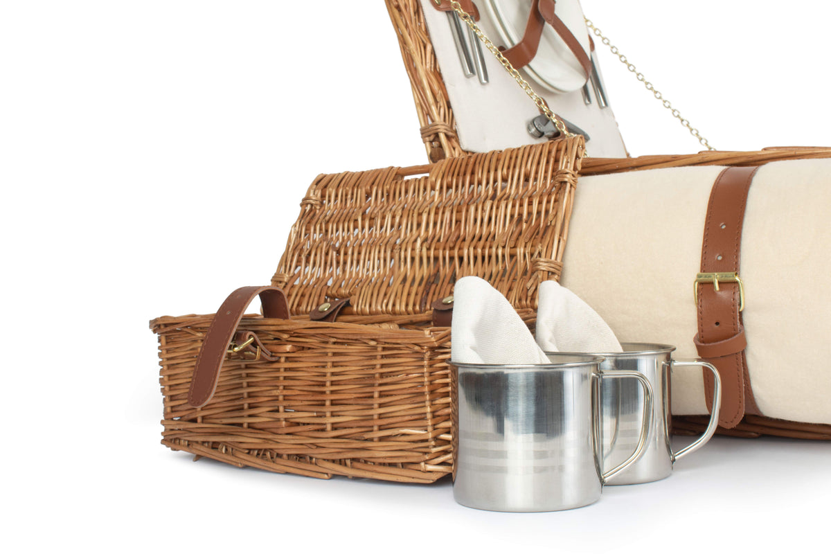 Open willow mug carrier with two stainless steel mugs beside the cream picnic rug from the Deluxe 2 Person Willow Picnic Hamper