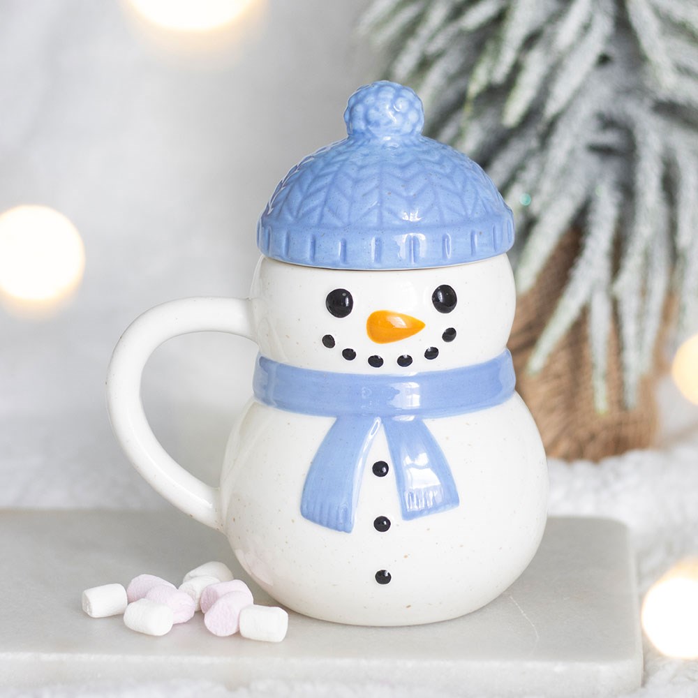 Winter Snowman Ceramic Mug with blue bobble hat lid and scarf displayed in a festive winter setting with marshmallows